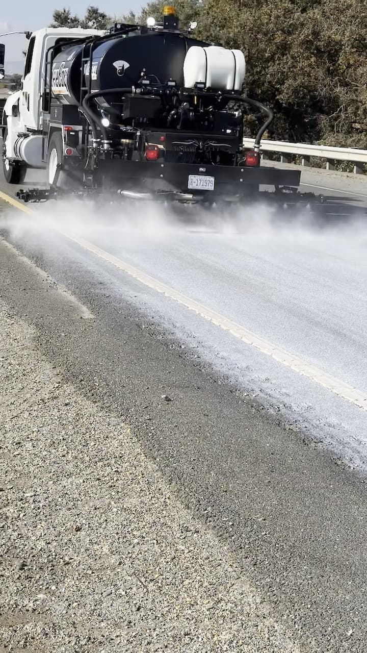 Distributor truck with two tanks spraying a surface treatment along a highway lane beside a guardrail