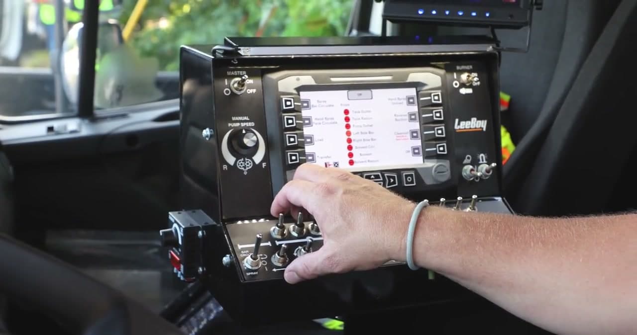 Operator’s hand at the in-cab control console of a LeeBoy PR900 distributor, a color display above a row of toggle switches